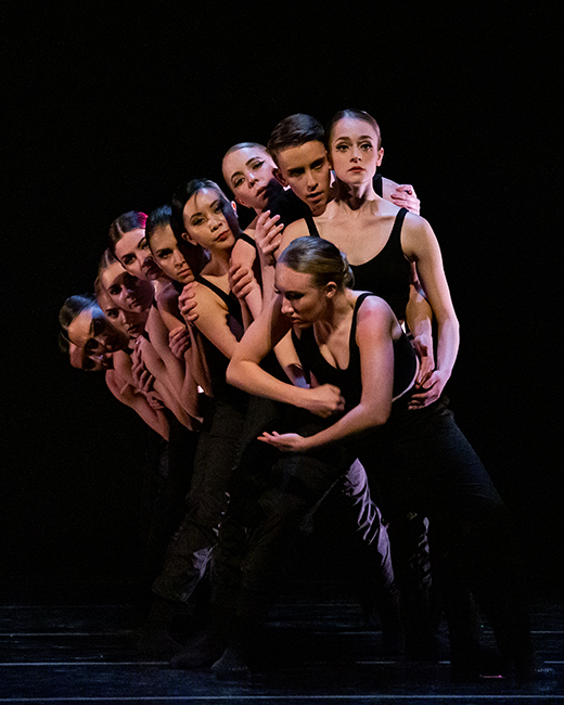 Group of pre-professional dance company dancers on stage huddled together looking over each others shoulders at audience